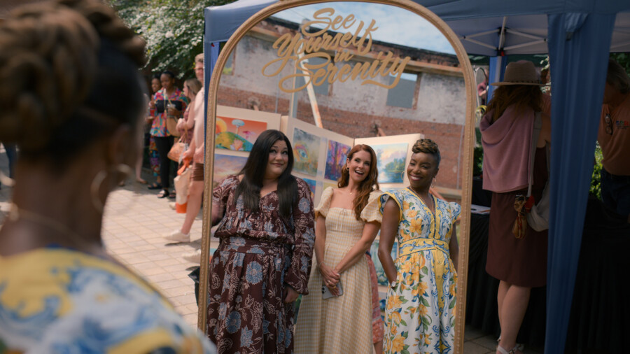 "Three women in colorful dresses smile together in front of a decorative mirror with "See yourself in serenity" at an outdoor event, surrounded by people and tents on a sunny day."