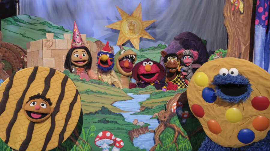 "Colorful puppets in costume pose in a whimsical fantasy landscape with a castle, bright sun, river, and giant cookies, evoking a playful, magical, and storybook setting reminiscent of a puppet show."