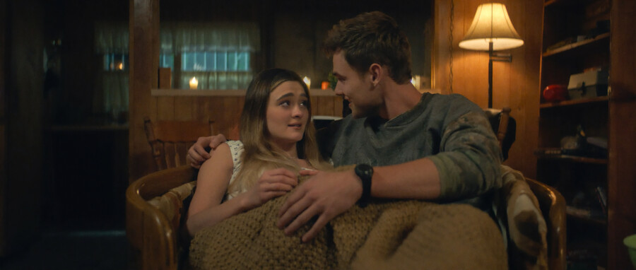"A young couple sits close together on a chair, wrapped in a blanket in a cozy, warmly lit wooden room with a lamp, shelves, and candles in the background, creating an intimate and comfortable atmosphere."
