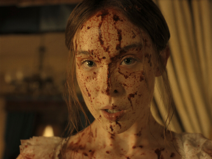 "A young woman with long hair, dressed in light clothing, has her face and upper chest covered in blood, standing indoors with dim warm lighting and blurred curtains and furnishings in the background."