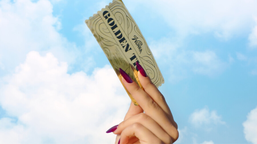 "A hand with long, painted nails holds up a golden ticket against a blue sky with clouds."