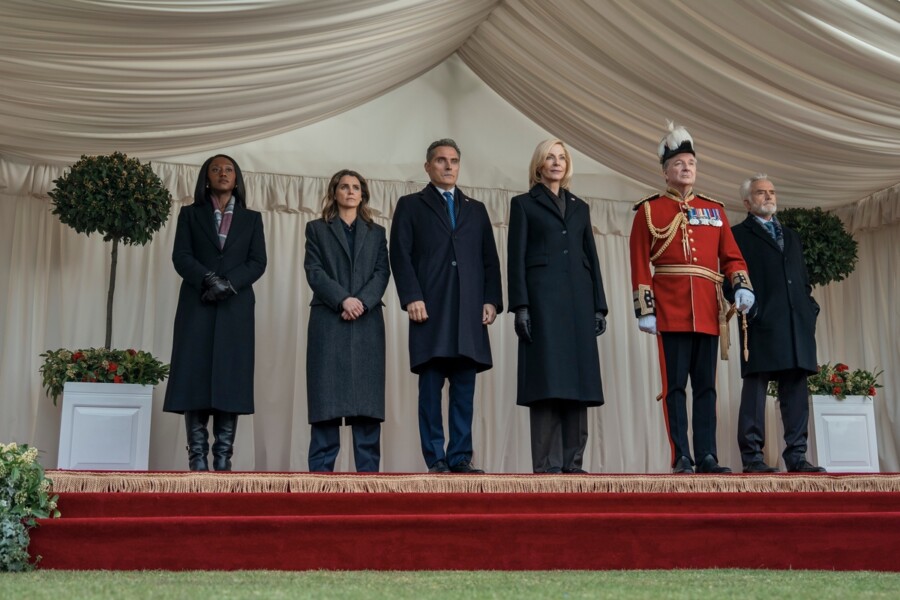 "Six formally dressed people stand on a red-carpeted stage under a draped white canopy, flanked by potted plants; one wears a ceremonial military uniform, the others wear dark coats, appearing to attend an official or formal outdoor event."
