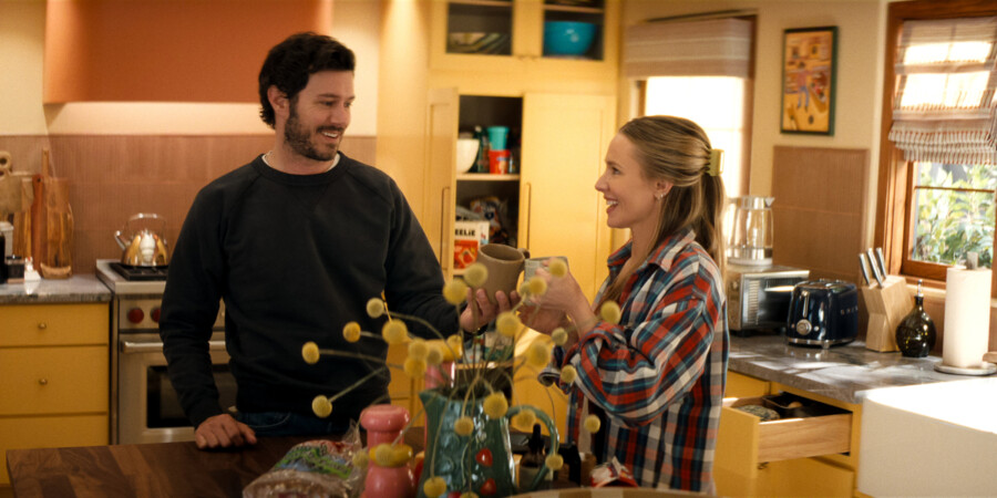 "A man and woman smile and toast with mugs in a cozy, sunlit kitchen, surrounded by yellow cabinets, kitchen appliances, and decorative items, creating a warm, homey atmosphere."